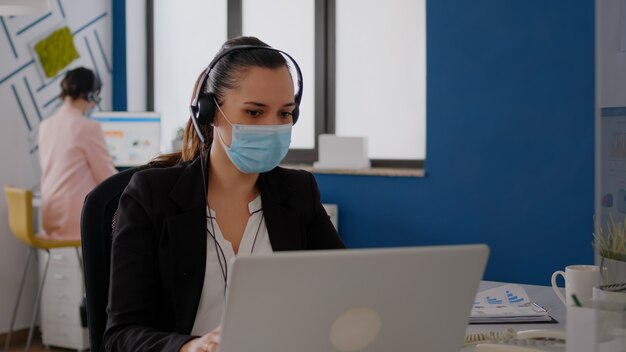 Executive Manager, der eine schützende Gesichtsmaske und einen Kopfhörer trägt, der mit dem Team in das Mikrofon spricht, das am Geschäftsprojekt arbeitet. Geschäftsfrau, die während der Coronavirus-Epidemie in einem neuen normalen Büro sitzt