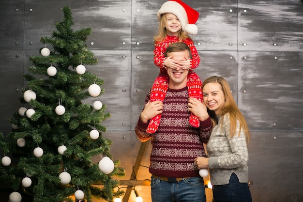 Kostenloses Foto familie durch den weihnachtsbaum mit kleiner tochter in einem roten hut