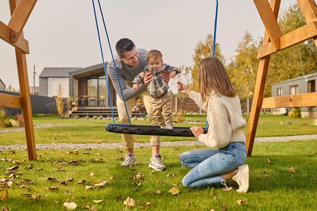 Familie, Freizeit. Junger erwachsener lächelnder mann, der kind auf schaukel stützt, und frau, die mit ausgestreckter hand hockt und am herbsttag zusammen in der nähe des landhauses ihre freizeit verbringt