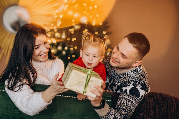 Familie mit der kleinen Tochter, die durch Weihnachtsbaum sitzt und Geschenkbox auspackt