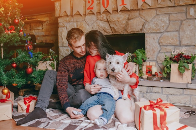 Familie vor dem Kamin sitzen mit ihrem Baby und ihrem Hund und einem Weihnachtsbaum