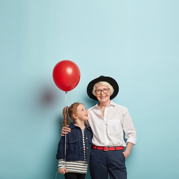 Familienfeier-Konzept. Nettes rothaariges Mädchen gratuliert reifer Oma mit Muttertag, hält roten Luftballon, umarmt zusammen, isoliert über blauer Wand mit Leerzeichen.