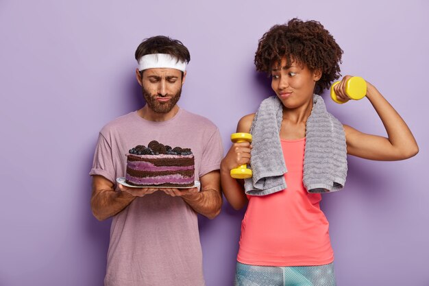 Familienpaar hat aktives Training, trägt Sportkleidung, hebt Hanteln, steht nebeneinander und schaut mit Versuchung auf Kuchen