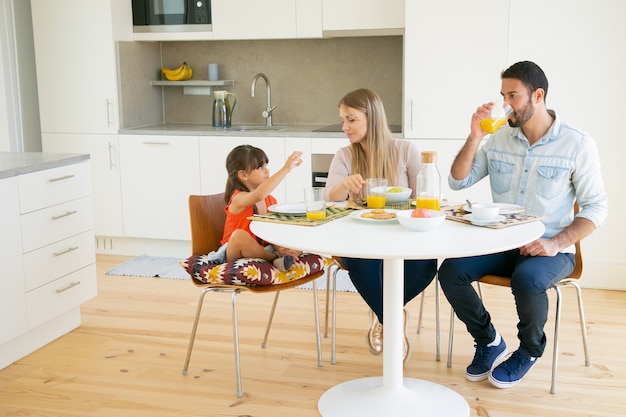 Familienpaar und Mädchen frühstücken zusammen in der Küche, sitzen am Esstisch, trinken Orangensaft und reden.
