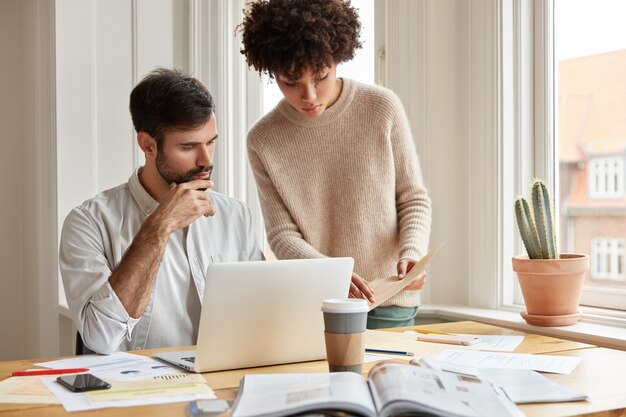 Familienpaare gemischter Abstammung untersuchen Papierkram, fragen sich gegenseitig um Rat, posieren gegen gemütliches Wohninterieur in der Nähe des Fensters, benutzen einen Laptop, trinken Kaffee zum Mitnehmen.