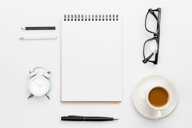 Farbige Bleistifte, Wecker, Stift, Brillen und gewundener Notizblock mit Kaffeetasse auf weißem Hintergrund