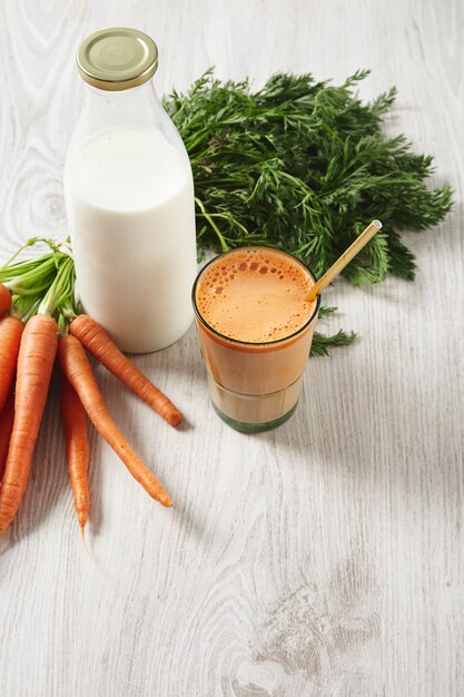 Farm Karottenernte in der Nähe von Milchflasche und Glas gefüllt mit Mischung aus natürlichem frischem Saft und Milch mit goldenem Trinkhalm darin