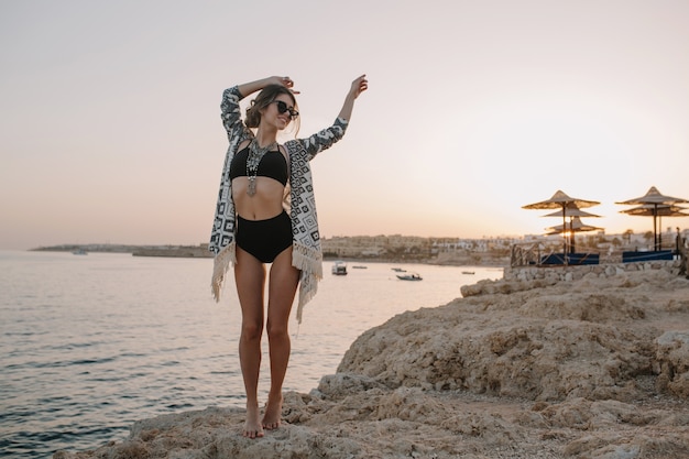 Fasionable Modell posiert am Strand, am Sonnenuntergang, Mädchen im Urlaub. Sexy Frau im schwarzen Bikini, Badeanzug mit hoher Taille, Strickjacke, Umhang mit Ornamenten.