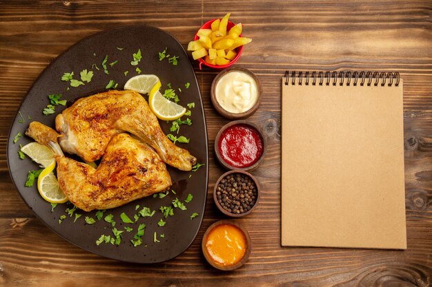 Fast Food von oben in den Tellerschüsseln mit schwarzen Pfeffersaucen und Pommes frites zwischen dem Teller mit Hähnchenschenkeln und Sahnenotizbuch