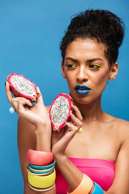 faszinierende Afrofrau mit bunten Kosmetika auf dem Gesicht, das auf der saftigen Drachefrucht schaut, schnitt zur Hälfte das Halten in beiden Händen, die, über blauer Wand lokalisiert wurden