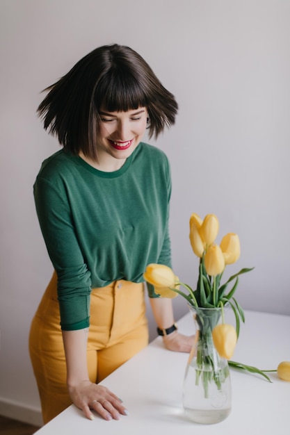 Faszinierendes brünettes Mädchen, das lacht, während es in der Nähe einer Blumenvase posiert Inspirierte kaukasische Dame, die Tulpen mit einem Lächeln betrachtet