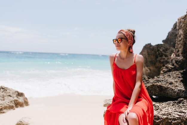 Faszinierendes weißes weibliches Modell, das Meerblick durch Sonnenbrille genießt. Foto im Freien der entspannten jungen Dame im roten Kleid, das auf Felsen nahe Meer aufwirft.