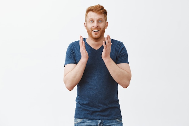 Kostenloses Foto fasziniert von wunderschöner landschaft. erstaunter und fröhlicher gutaussehender rothaariger mann mit borsten im blauen t-shirt, erhobene handflächen, die erfreut und aufgeregt blicken und über der grauen wand unter eindruck stehen