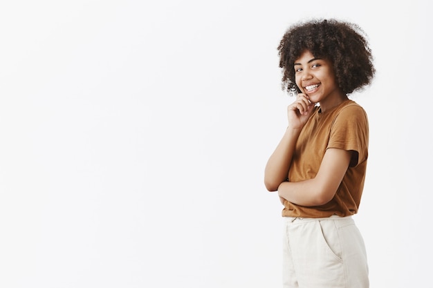 Faszinierte flirty weibliche dunkelhäutige Frau mit Afro-Frisur in braunem T-Shirt, das im Profil steht und sich mit niedlichem Lächeln dreht, das Hand auf Kinn hält, das mit Interesse und Verlangen schaut