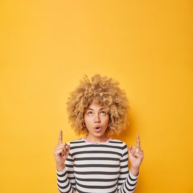 Kostenloses Foto faszinierte lockige frau mit lockigen haarpunkten über beeindruckt zeigt erstaunliche einkaufsangebote oder werbung trägt einen lässigen gestreiften pullover, der über gelbem hintergrund isoliert ist, hat ein überraschtes gesicht