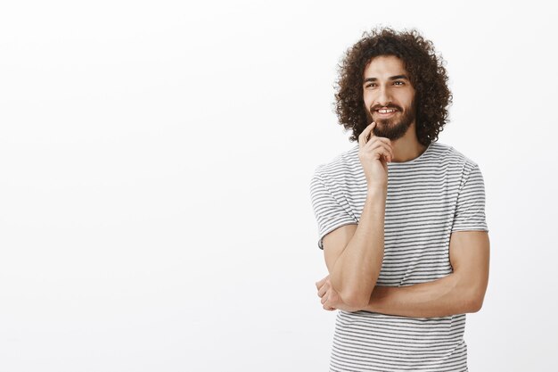 Faszinierter fröhlicher bärtiger Mann mit Afro-Frisur und Bart, der zur Seite schaut, den Finger auf der Lippe hält und neugierig lächelt und über etwas Leckeres oder Interessantes nachdenkt