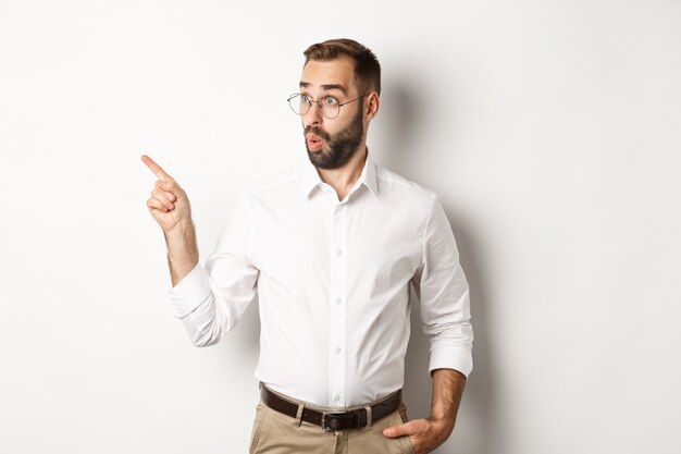 Faszinierter junger Mann in der Brille zeigt mit dem Finger nach links und betrachtet das interessierte Promo-Angebot stehend