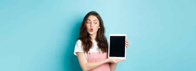 Fasziniertes Glamour-Girl, das auf den Bildschirm des digitalen Tablets schaut, während es Ihnen amüsiert über das Online-Promo-Angebot steht, demonstriert etwas auf dem blauen Hintergrund