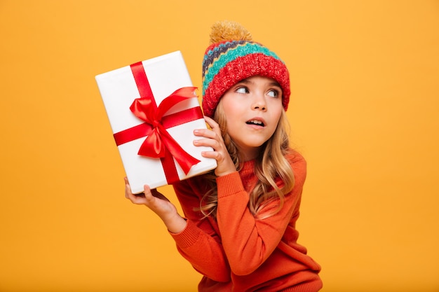 Fasziniertes junges Mädchen in der Strickjacke und in Hut, die Geschenkbox halten und weg über Orange schauen