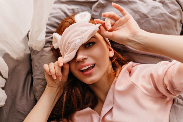 Faule rothaarige Frau, die in Schlafmaske posiert Overhead-Foto eines neugierigen rothaarigen Mädchens, das im Bett liegt