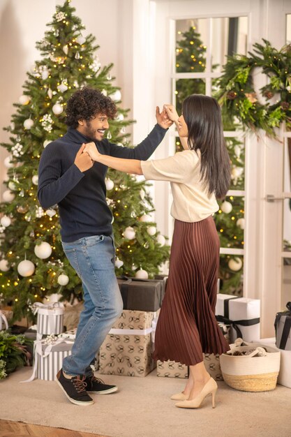 Feier. Junger bärtiger lächelnder indischer Mann und hübsche asiatische Frau in High Heels, die sich gegenseitig tanzende berührende Hand in der Nähe von Weihnachtsbaum und Geschenken betrachten