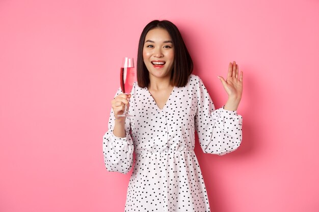 Feier. Schöne asiatische Frau, die Champagner trinkt und lächelt, im Kleid über rosa Hintergrund stehend.