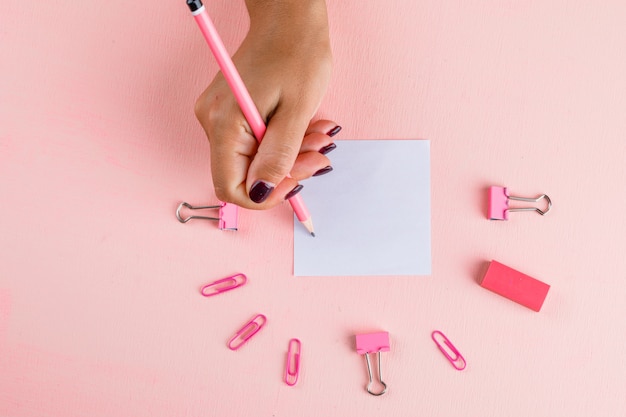 Feierkonzept mit Papier- und Binderclips, Radiergummi auf rosa Tisch flach legen. Frau, die auf Haftnotiz schreibt.