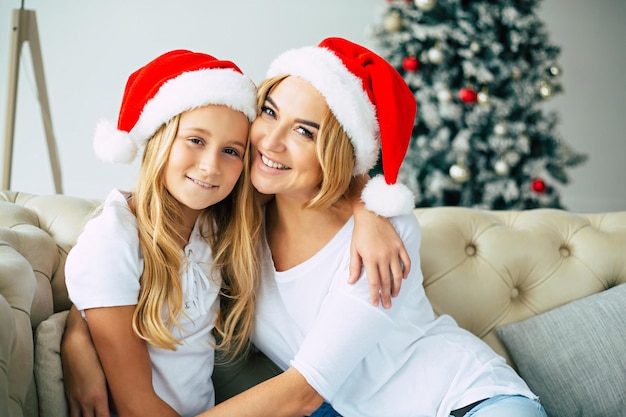 Feiern, feiertage, familie, weihnachten, neujahr! schöne glückliche mutter und ihre kleine tochter in weihnachtsmützen sitzen und umarmen sich auf weihnachtsbaumhintergrund