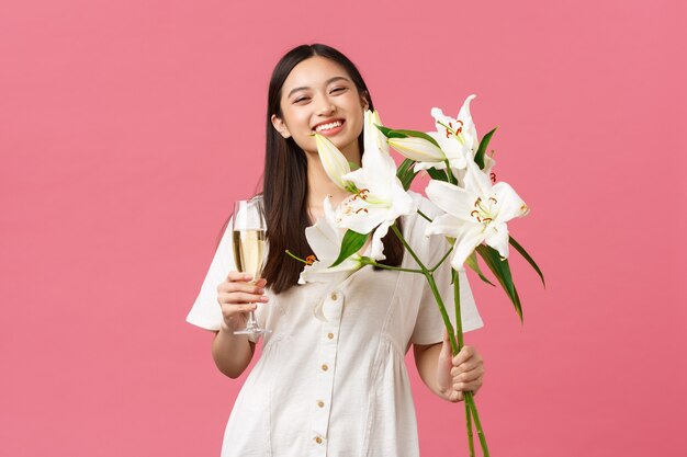 Feiern, Partyferien und lustiges Konzept. Dummes alles Gute zum Geburtstagsmädchen im weißen Kleid, breit lächelnd, während es einen schönen Blumenstrauß aus Lilien erhält, ein Glas Champagner hält und rosa Hintergrund steht