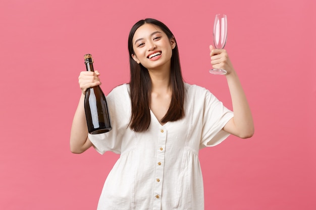 Feiern, Partyferien und lustiges Konzept. Fröhliches, glückliches asiatisches Mädchen, das bereit ist, den freien Tag mit Freundinnen zu genießen, Champagner und Gläser mitzubringen, lächelnde Kamera, fröhlicher rosa Hintergrund.