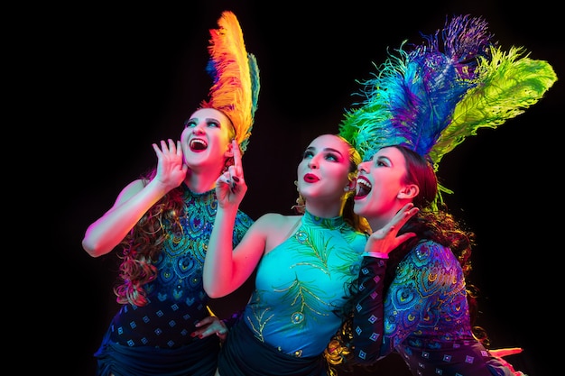 Feiern. Schöne junge Frauen im Karneval, stilvolles Maskenkostüm mit Federn auf schwarzem Hintergrund im Neonlicht. Exemplar für Anzeige. Feiertagsfeier, Tanzen, Mode. Festliche Zeit, Party.