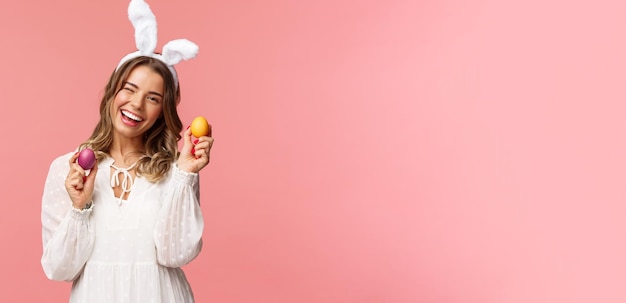 Feiertage Frühlings- und Partykonzept Fröhliche, gutaussehende blonde Frau, die den Ostertag in Hasenohren feiert und zwei bemalte Eier und eine Zwinkerkamera hält, die glücklich auf rosafarbenem Hintergrund lächelt