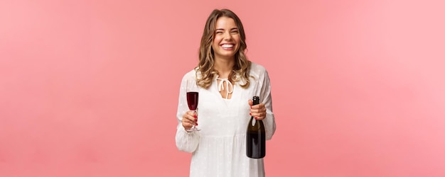 Feiertage Frühlings- und Partykonzept Porträt einer glücklichen und sorglosen europäischen blonden Frau, die in weißem Kleid feiert und eine Flasche Champagner oder Wein aus Glas trinkt und lacht