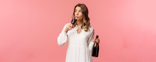 Feiertage Frühlings- und Partykonzept Porträt einer sorglosen, unabhängigen, süßen, blonden Frau, die Wein aus einer Glasflasche trinkt und ein Getränk verkostet, schauen Sie nach oben und feiern Sie mit Freunden auf rosafarbenem Hintergrund