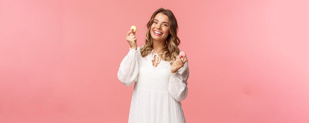 Feiertage Frühlings- und Partykonzept Porträt einer zarten, schönen blonden Frau in weißem Kleid, die fröhlich mit zwei Macarons tanzt, die glücklich lächeln und köstliches Dessert schmackhaftes Essen auf rosa Hintergrund essen