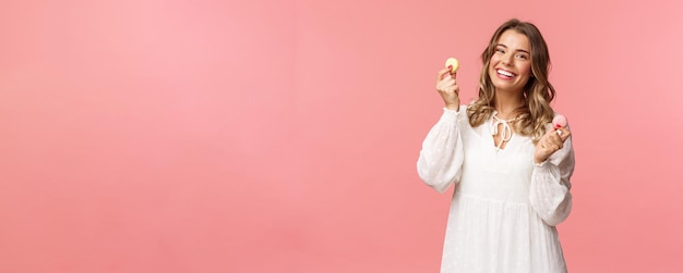 Feiertage Frühlings- und Partykonzept Porträt eines süßen romantischen blonden Mädchens in weißem Kleid, das die Lippen leckt, als verlockend, ein köstliches Dessert mit zwei Macarons zu essen und einen zufriedenen rosa Hintergrund zu sehen