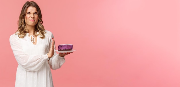 Feiertage Frühlings- und Partykonzept Porträt eines unzufriedenen und unzufriedenen blonden Mädchens in weißem Kleid, das ablehnendes Essen von geschmacklosem Kuchen mit Grimassen ablehnt und Stoppschild auf dem rosafarbenen Hintergrund des Desserts zeigt