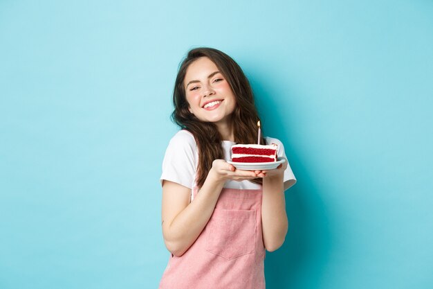 Feiertage und Feiern. Nettes Glamour-Mädchen, das ihren Geburtstag feiert, Teller mit Kuchen hält und fröhlich lächelt, feiert, auf blauem Hintergrund steht.