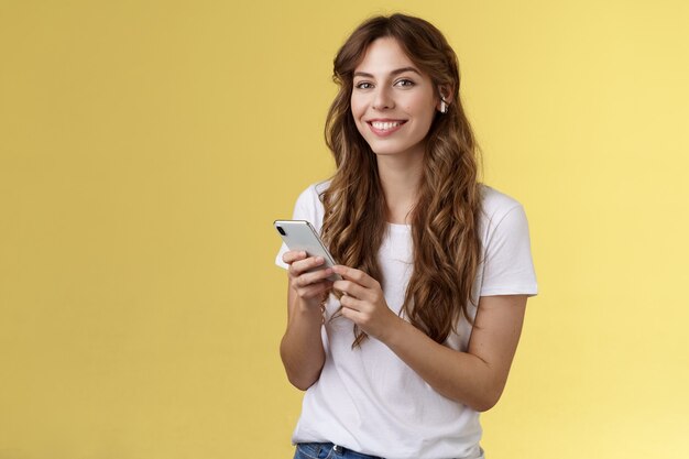 Feminine schöne lockige Frau weißes T-Shirt halten Smartphone ausgewählt tolles neues Lied hören Musik mit drahtlosen Kopfhörern lächelnd erfreut Kamera genießen Ohrhörer schlägt gelben Hintergrund