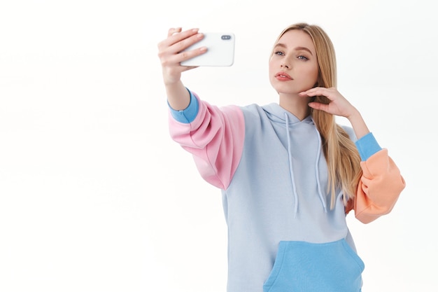 Feminines, gut aussehendes blondes Mädchen mit sinnlichem Ausdruck, das das Gesicht sanft berührt, als es ein Selfie auf dem Handy macht