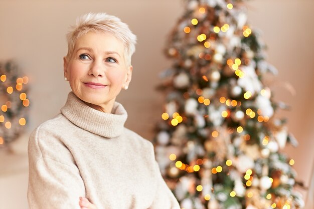 Ferienzeit, Tradition und Feierkonzept. Attraktive sechzigjährige Frau im kuscheligen Pullover, der im Wohnzimmer steht, verziert mit majestätischem Weihnachtsbaum mit Ornamenten, Girlanden und Lichtern