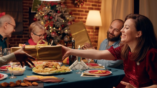 Festliche Familienmitglieder sitzen am Weihnachtstisch und tauschen Geschenke zu Hause aus. Fröhliche Menschen versammelten sich im Wohnzimmer, um den traditionellen Winterurlaub zu feiern und sich gegenseitig Geschenke zu machen.