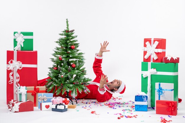 Festliche Feiertagsstimmung mit jungem nervösem Weihnachtsmann, der hinter Weihnachtsbaum nahe Geschenken auf weißem Hintergrund liegt