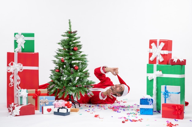 Festliche Feiertagsstimmung mit jungem Weihnachtsmann, der hinter Weihnachtsbaum nahe Geschenken auf weißem Hintergrund lagert Foto auf Lager