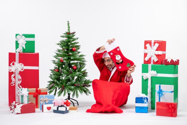 Festliche Feiertagsstimmung mit lustigem positiv überraschtem Weihnachtsmann, der auf dem Boden sitzt und Weihnachtssocke nahe Geschenken und geschmücktem Weihnachtsbaum auf weißem Hintergrund zeigt