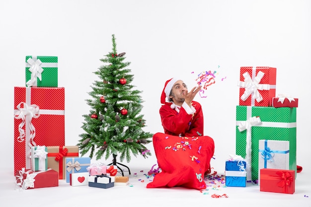 Festliche Feiertagsstimmung mit lustigem Weihnachtsmann, der auf dem Boden sitzt und mit Weihnachtsdekorationen nahe Geschenken und geschmücktem Weihnachtsbaum auf weißem Hintergrund spielt