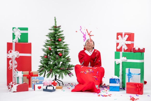 Festliche Feiertagsstimmung mit positivem Weihnachtsmann, der auf dem Boden sitzt und mit Weihnachtsdekorationen nahe Geschenken und geschmücktem Weihnachtsbaum auf weißem Hintergrund spielt