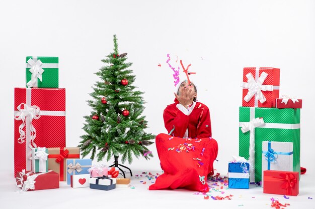 Festliche Feiertagsstimmung mit positivem Weihnachtsmann, der auf dem Boden sitzt und mit Weihnachtsdekorationen nahe Geschenken und geschmücktem Weihnachtsbaum auf weißem Hintergrund spielt