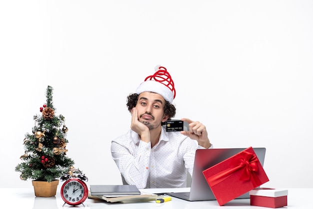 Festliche Stimmung des Feiertags mit dem jungen bärtigen Geschäftsmann mit Weihnachtsmannhut, der Bankkarte hält und seine Hand unter Kinn im Büro legt