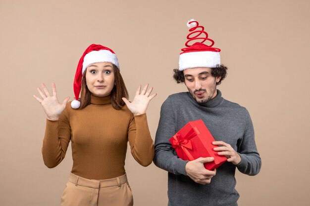 Festliches Konzept der Neujahrsstimmung mit dem lustigen glücklichen reizenden Paar, das rote Weihnachtsmannhüte auf grauem Lagerbild trägt